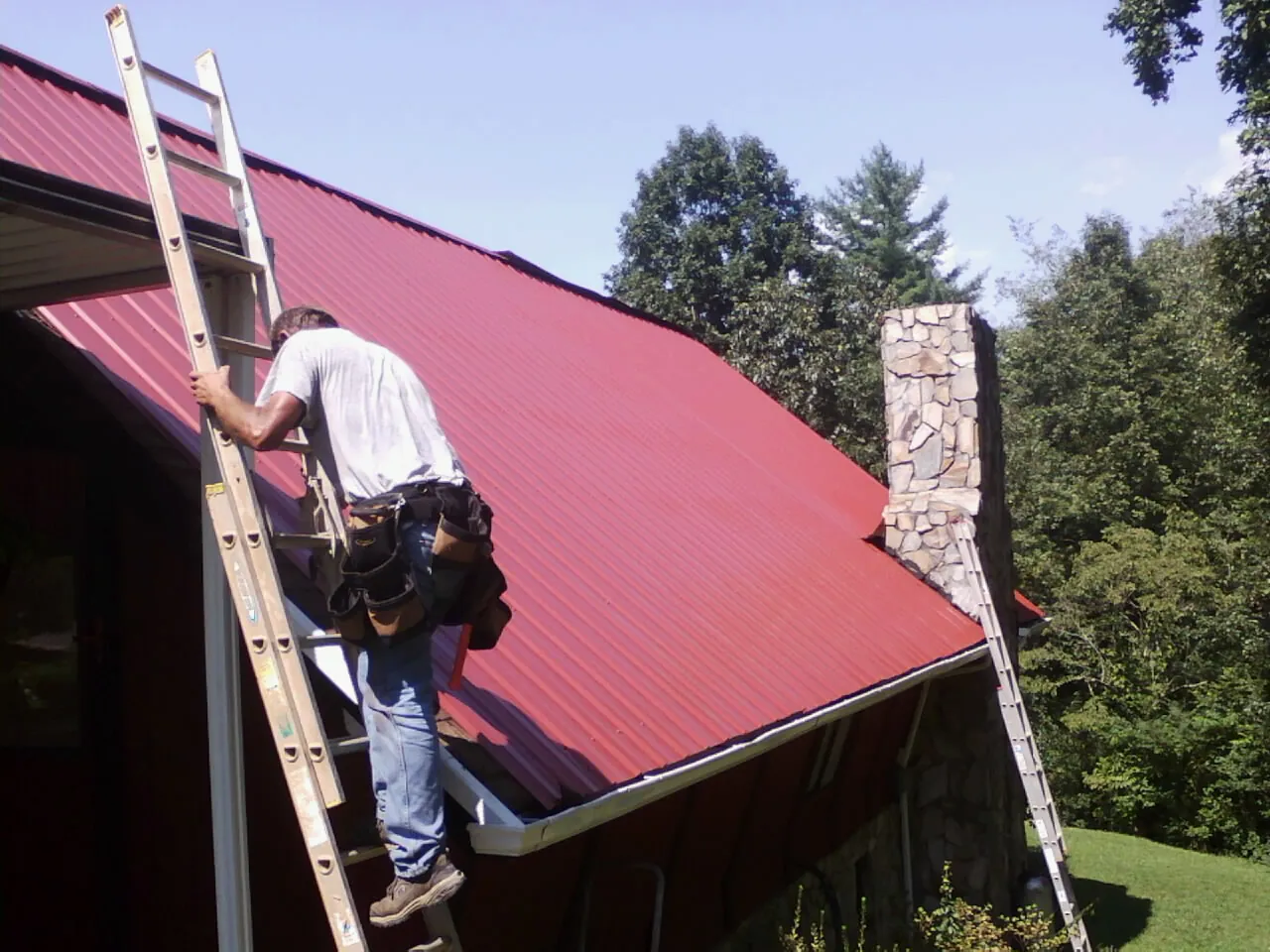 Roofer on a ladder beside a red metal roof and stone chimney — Soffit & Fascia Repair by Roofing Brothers in Emporia
